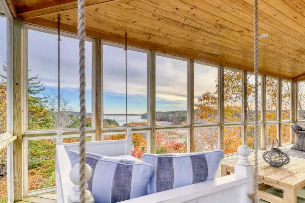 Porch swing with blue pillows on a screened porch, overlooking autumn trees and a lake in the distance.