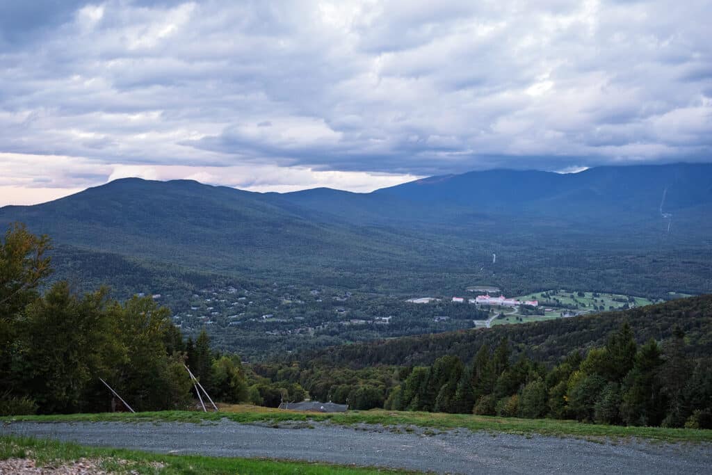 Mountain view with green forests, distant buildings, and a cloudy sky above a sloping gravel path.
