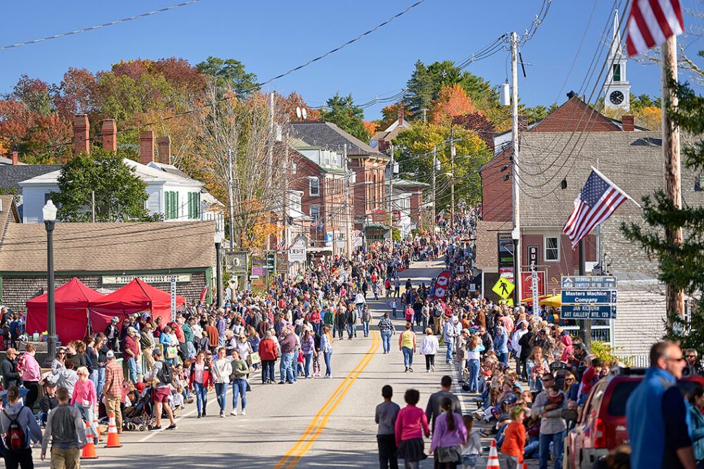 Crowds fill a small town street lined with buildings and autumn trees during a sunny festival or parade.