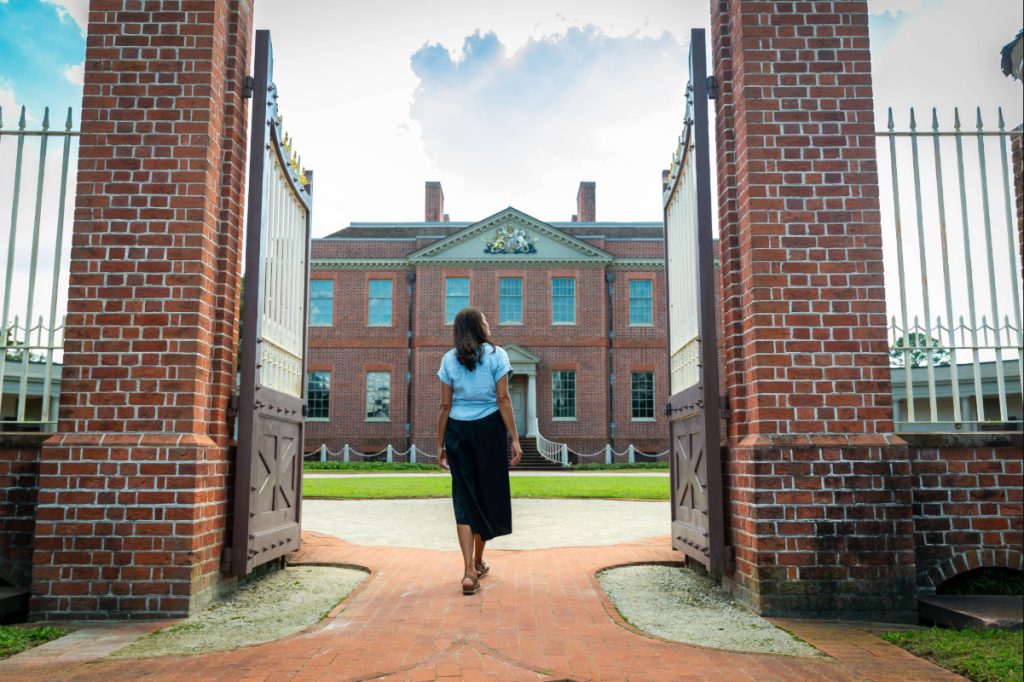 A woman walks through open brick gates toward a large historic brick building, Tryon Palace, in New Bern, North Carolina.