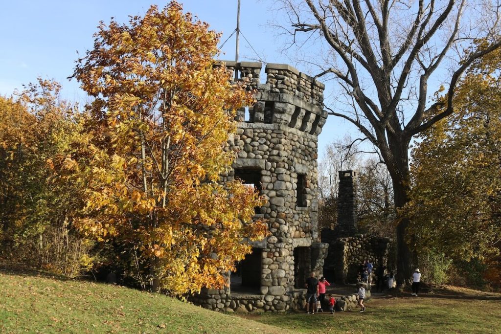 Stone tower surrounded by autumn trees; people gathered around the base on a sunny day.