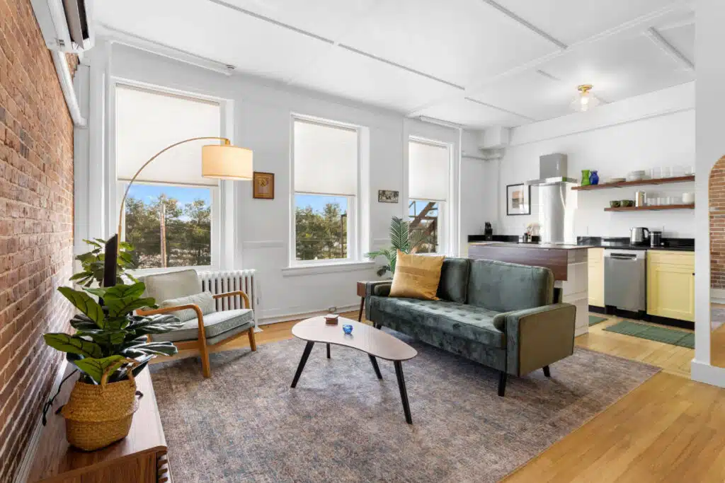 Rockland Maine apartment rental from Tides to Pines. Bright living room with a green sofa, armchair, plants, and large windows near an open kitchen.