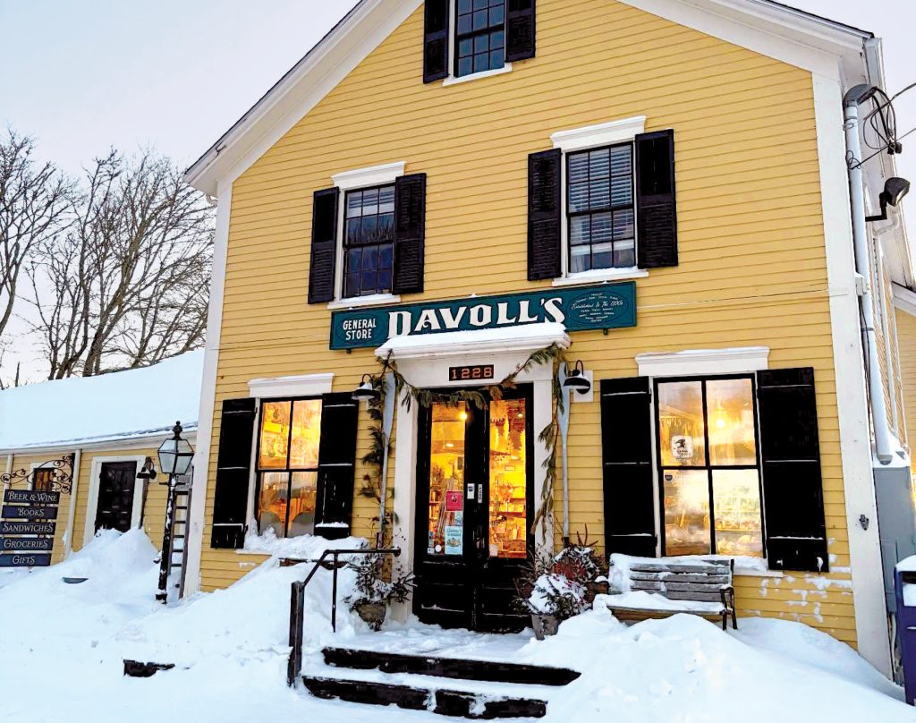 A yellow general store with black shutters sits in the snow, warmly lit from inside.