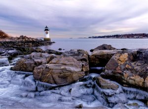 A lighthouse by the sea, surrounded by rocky, ice-covered shoreline under a cloudy sky.