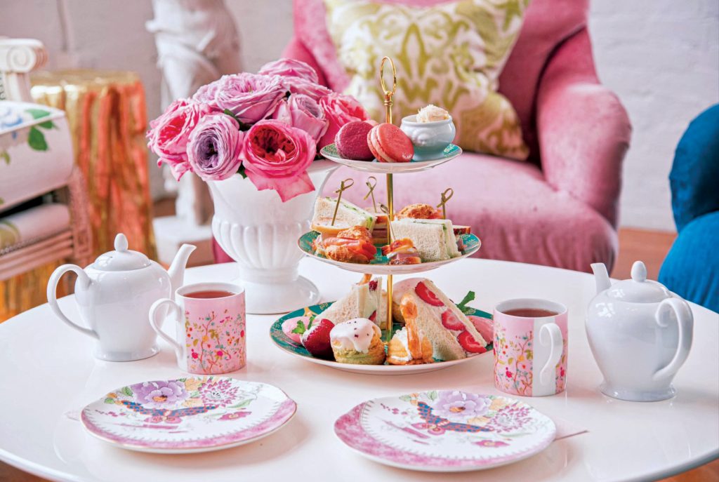 Afternoon tea set with pastries, sandwiches, floral plates, mugs, teapots, and pink roses on a white table.