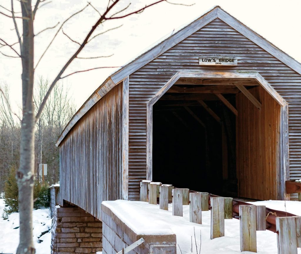 A wooden covered bridge crosses a snowy riverbank under a pale sky, surrounded by bare trees in winter.
