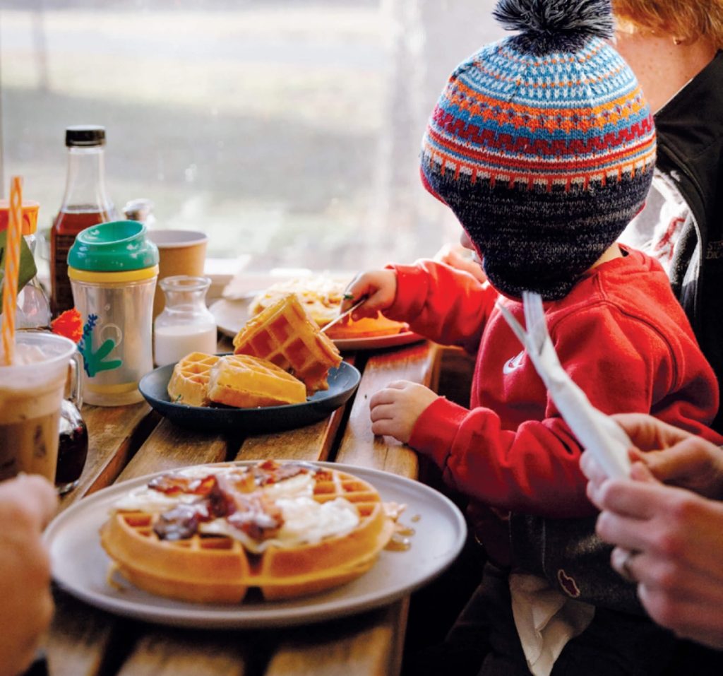 A child in a colorful hat eats waffles at a table with adults, drinks, and more food.