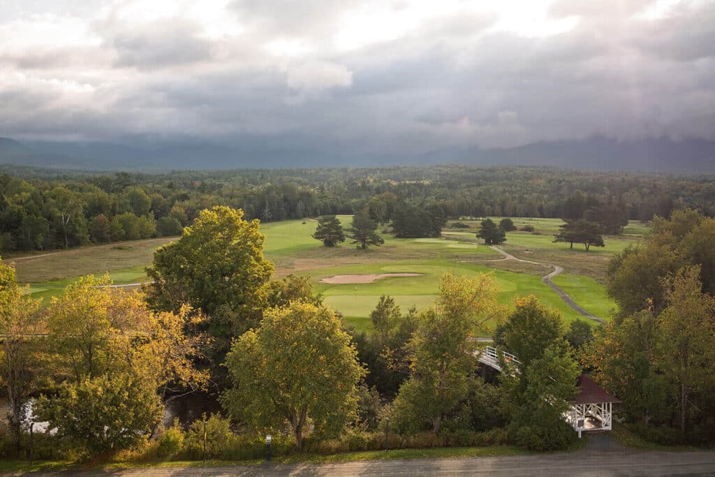 A green golf course surrounded by trees under a cloudy sky, with a small white gazebo in the corner.