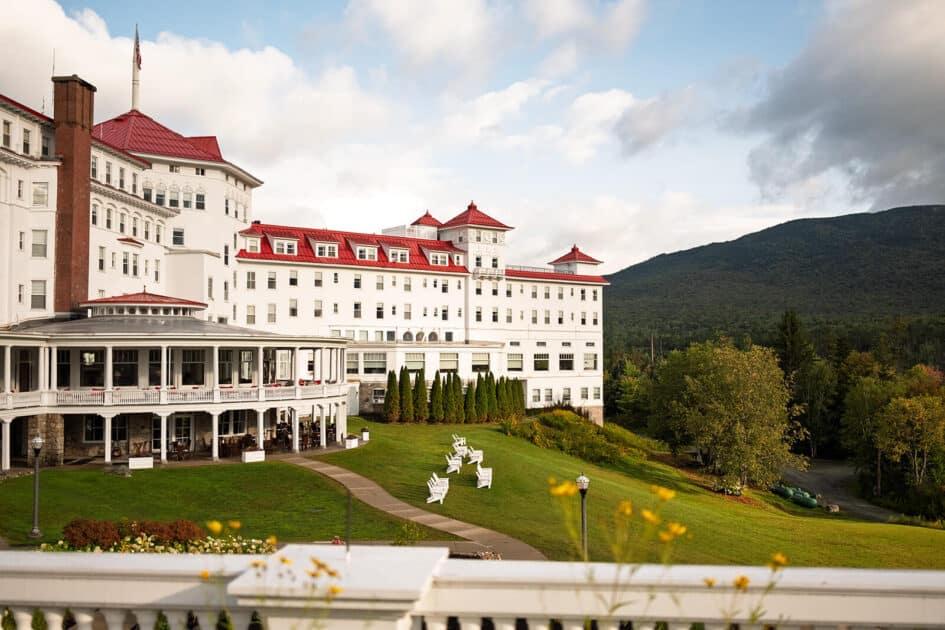 Large white hotel with red roofs, green lawn, and mountain backdrop under a partly cloudy sky.
