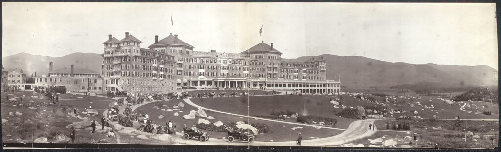 Large historic hotel with many cars parked outside, surrounded by hills and open land, circa early 1900s.