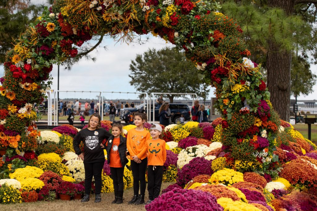 Four kids in orange and black stand under a large colorful mum flower arch at MumFest in New Bern.