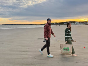 Two people walk on a sandy beach at sunset, carrying fishing gear and a Renys shopping bag.