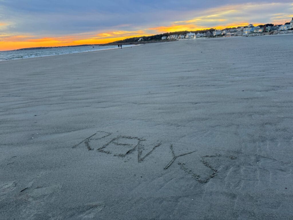 RENYS written in sand on a Maine beach at sunset, with buildings and people in the distance.
