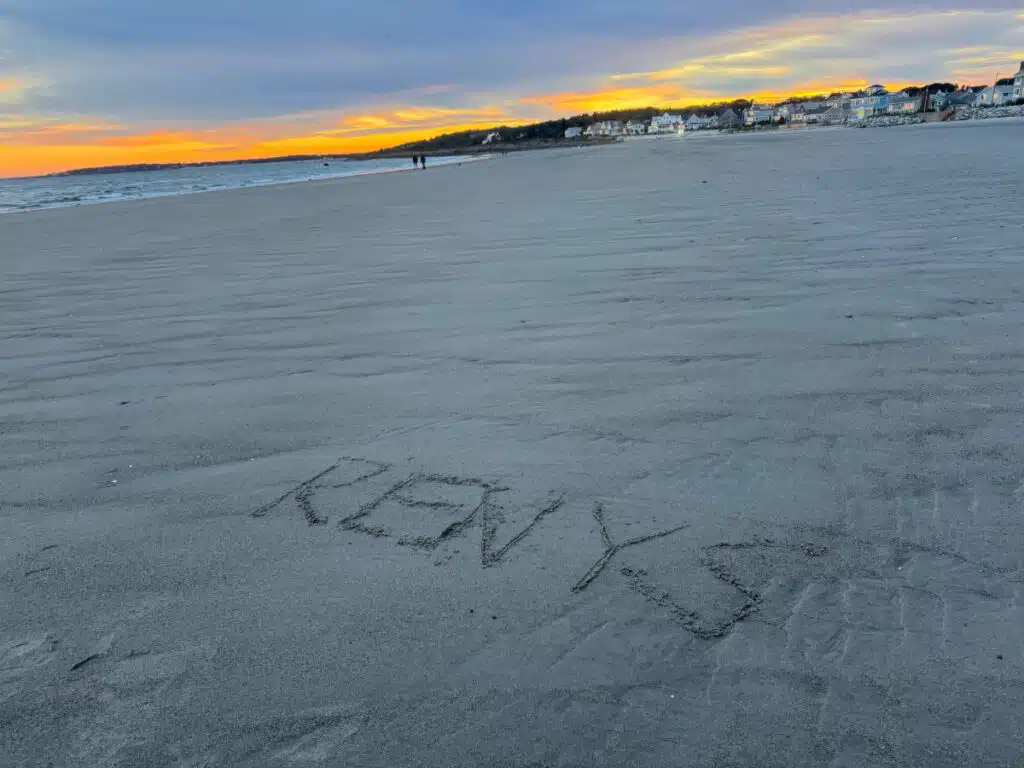 RENYS written in sand on a Maine beach at sunset, with buildings and people in the distance.