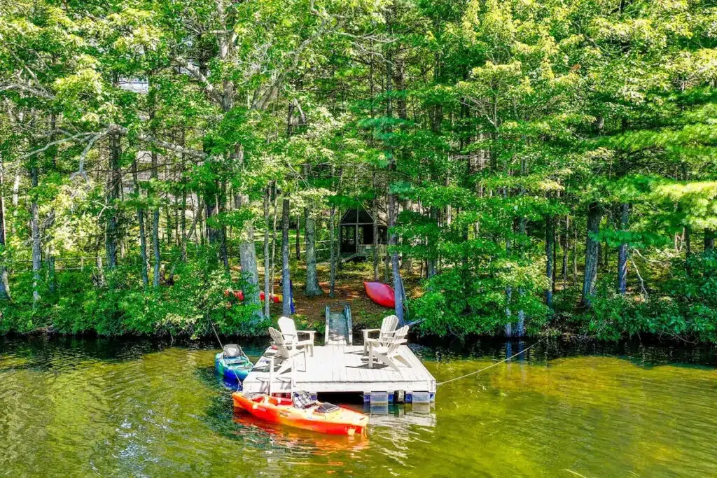 Maine vacation home with small wooden dock with chairs and kayaks, surrounded by trees on a calm, clear lake.