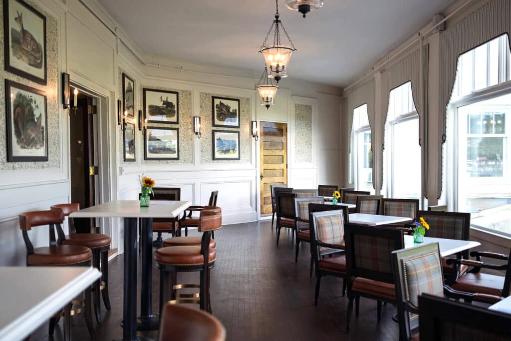 Bright cafe with large windows, framed animal art, wooden tables, and mixed plaid and leather chairs.