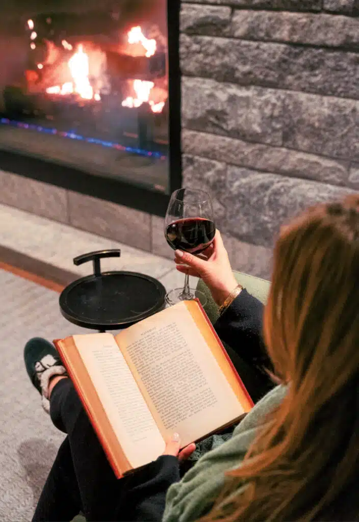 Person reading a book by a fireplace at the Williams Inn in Williamstown, holding a glass of red wine, seated in a cozy setting.