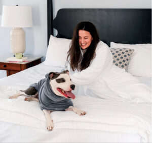 At the Williams Inn in Williamstown, Massachusetts, a woman in a white robe smiles on a bed next to a dog wearing a gray hoodie.