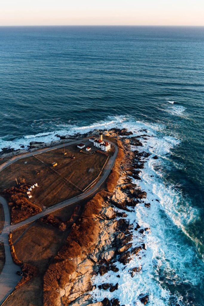 A lighthouse stands on a rocky coast, surrounded by waves, under a clear sky at sunset.