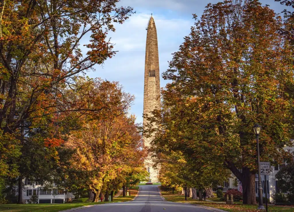 Tall stone obelisk surrounded by autumn trees along a quiet street under a partly cloudy sky.