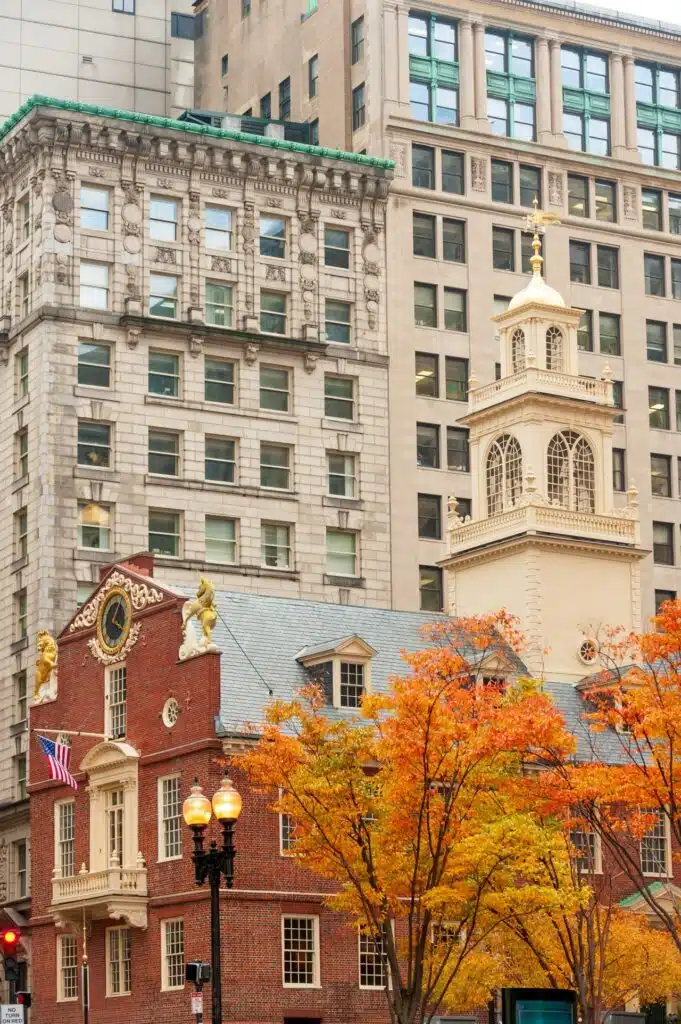 6 New England Places to Visit This Fall for America 250. Red-brick historic building with a clock, autumn trees, and tall modern buildings in the background.