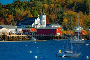 Sailboats on blue water, red buildings, and a white church with a steeple among autumn trees.