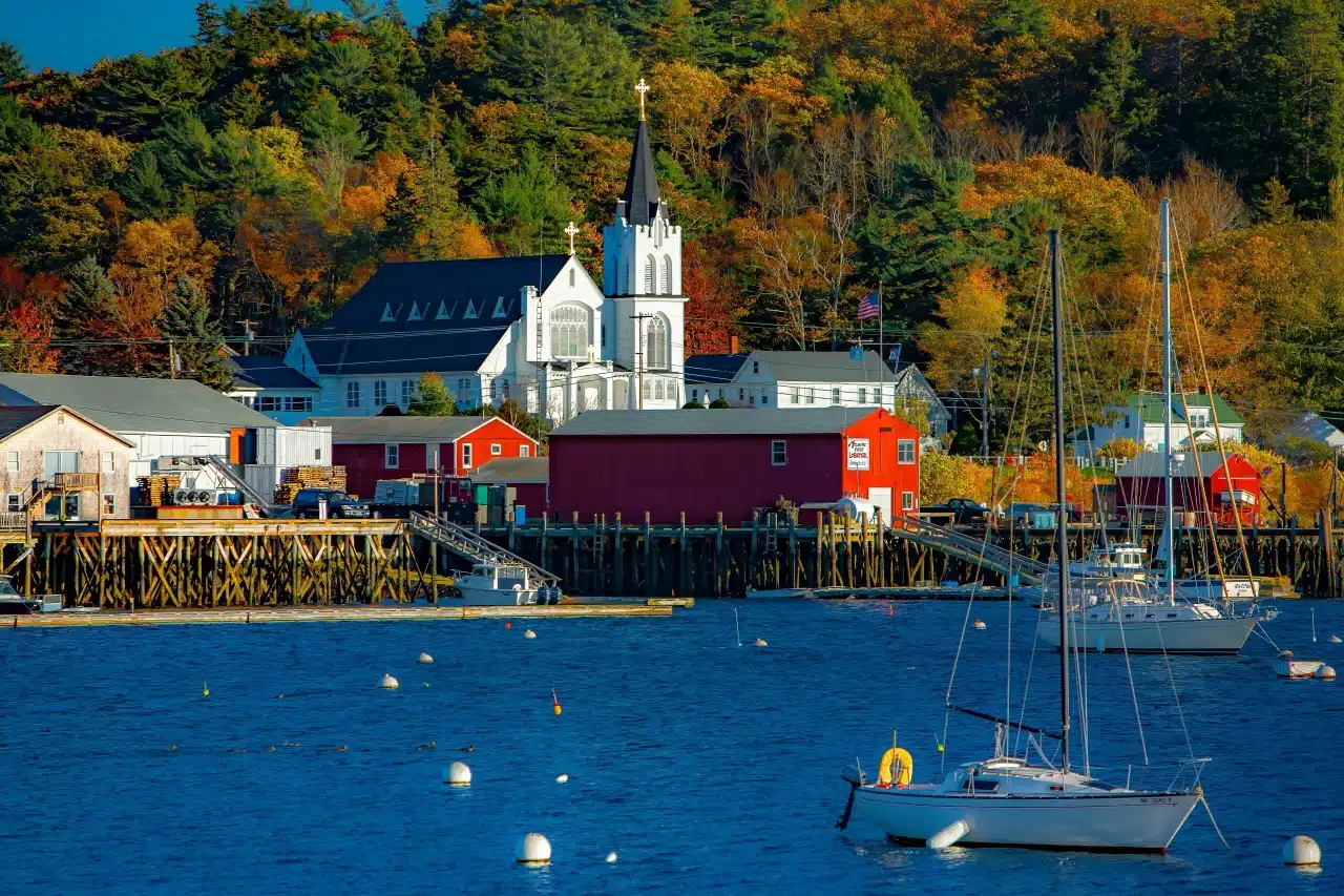 Sailboats on blue water, red buildings, and a white church with a steeple among autumn trees.