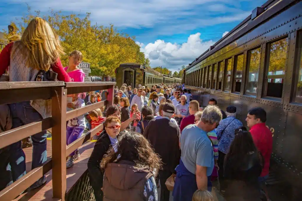 5 New England Coastal Towns to Visit for Fall Foliage. A crowd of people waits to board a vintage train on a sunny day, surrounded by trees with autumn leaves.