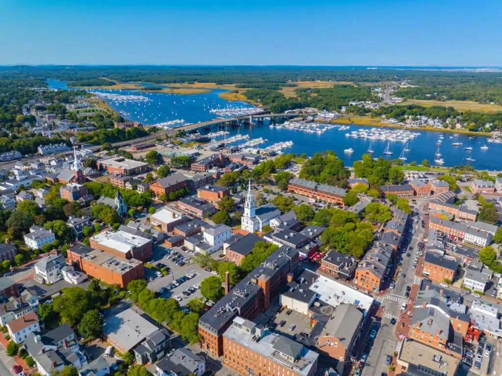 5 New England Coastal Towns to Visit for Fall Foliage. Aerial view of a coastal town with boats, a river, and historic brick buildings under a clear blue sky.