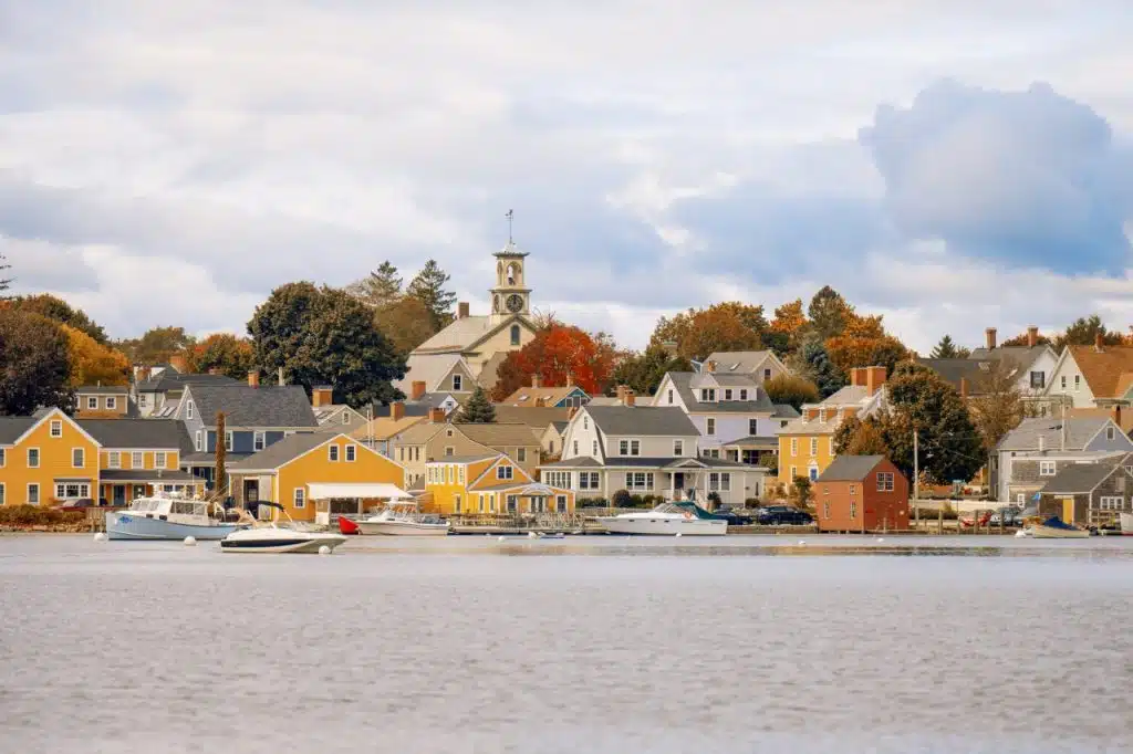 Boats docked by colorful houses and a church on a calm waterfront under a partly cloudy sky.