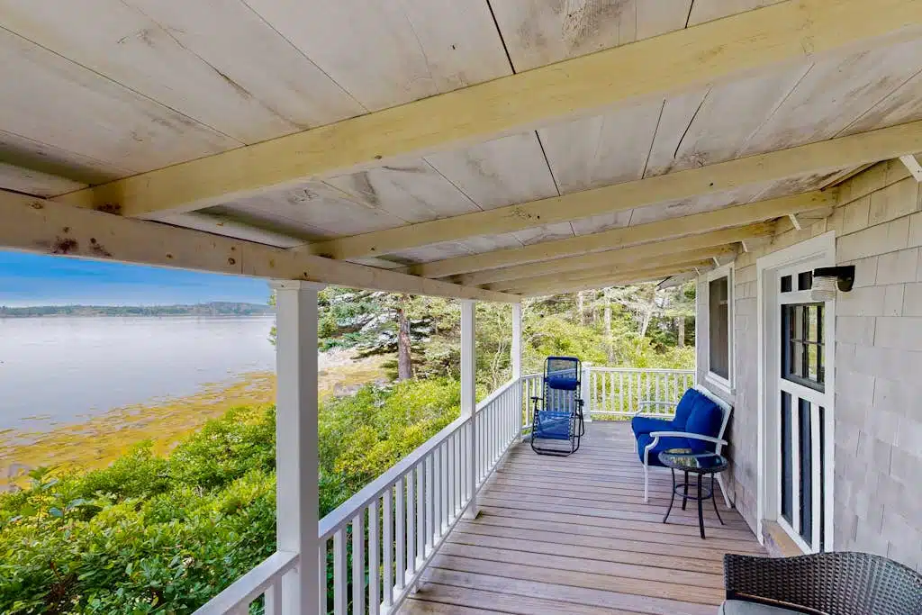 Covered porch with blue chairs overlooks water and trees, with white railing and wooden floor.