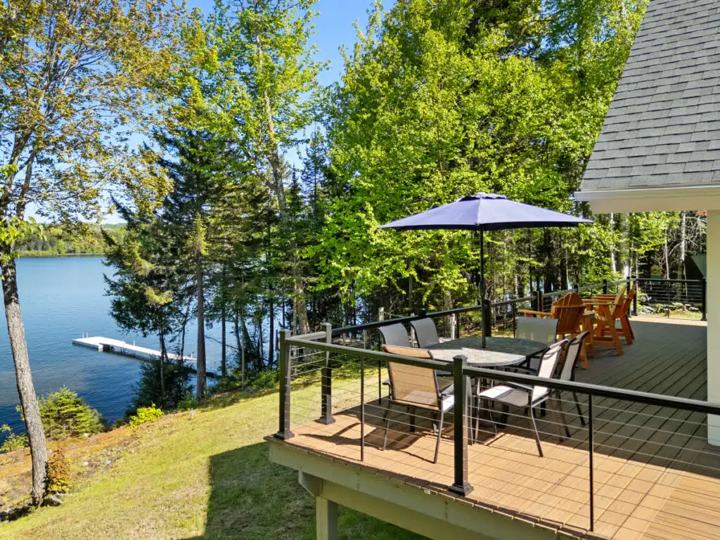 A lakeside Maine house deck with a table, chairs, umbrella, and a view of the water and dock.