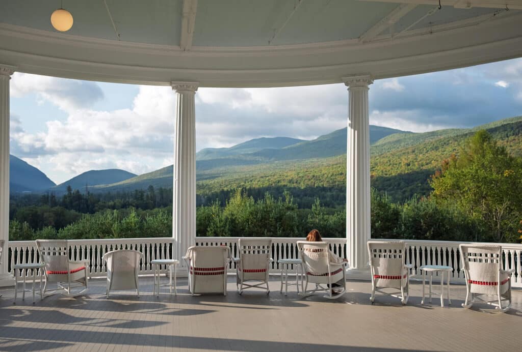 Person sits on a large porch with white chairs, overlooking green mountains and trees under a cloudy sky.