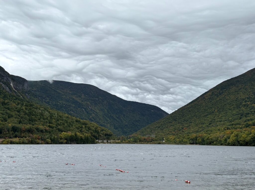 Cloudy sky over a lake, surrounded by forested mountains, with red buoys floating on the water.