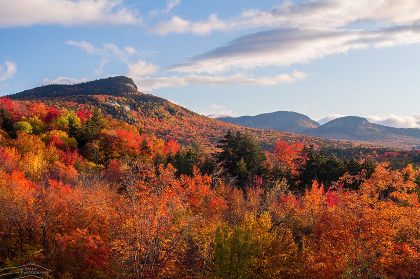 Colorful autumn trees cover rolling hills under a partly cloudy sky with distant mountains in the background.
