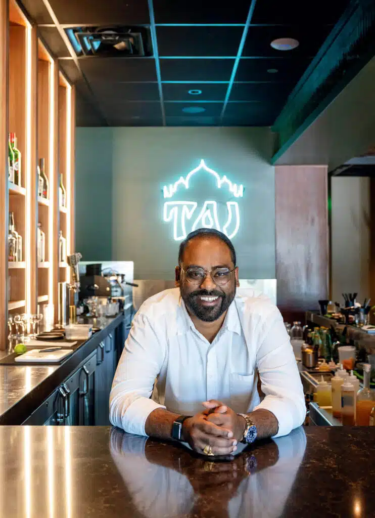 A smiling man in a white shirt leaning over a bar in front of a glowing neon Taj sign.