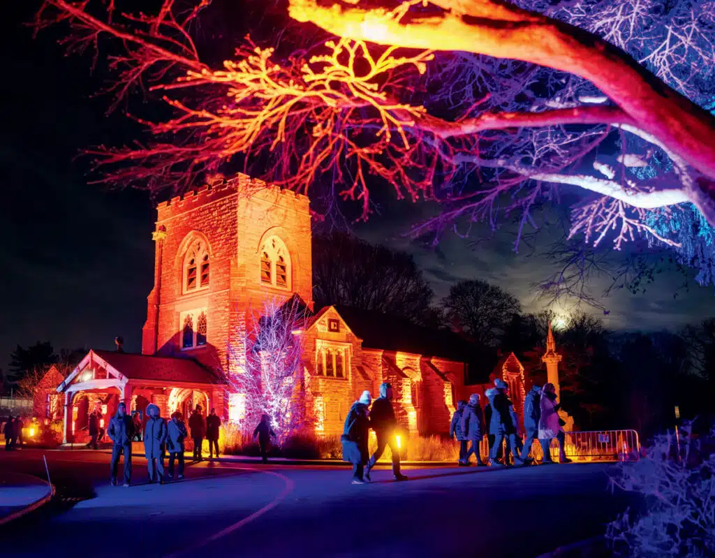 People walk toward a stone church illuminated by vibrant red and blue lights at night.