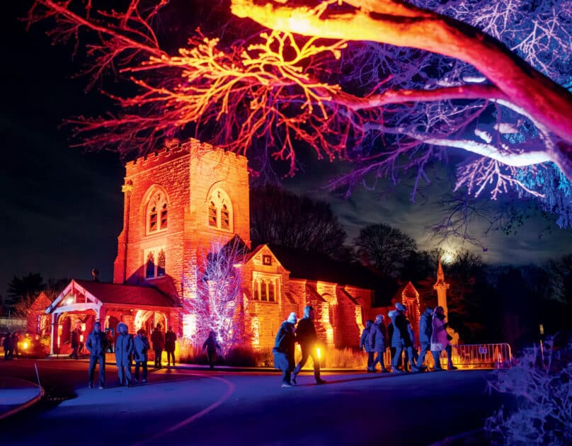 People walk toward a stone church illuminated by vibrant red and blue lights at night.