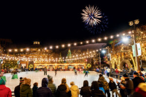 People ice skating at an outdoor rink at night with fireworks in the sky above.
