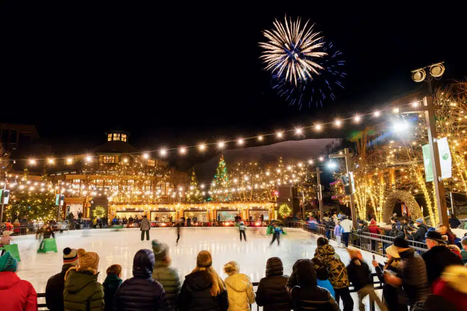 People ice skating at an outdoor rink at night with fireworks in the sky above.