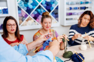 A person demonstrating knitting techniques to two women in a craft studio.