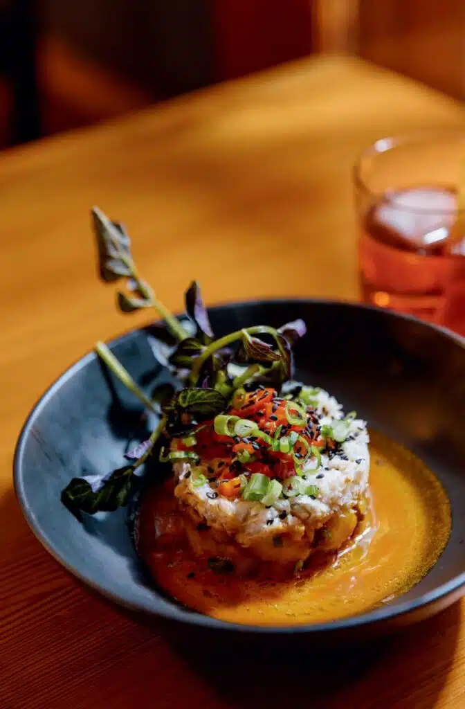 Crab cake topped with chopped vegetables and herbs in a shallow bowl of sauce.
