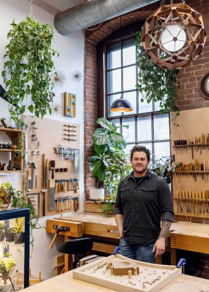 A woodworker stands in his sunlit workshop filled with tools and hanging plants.