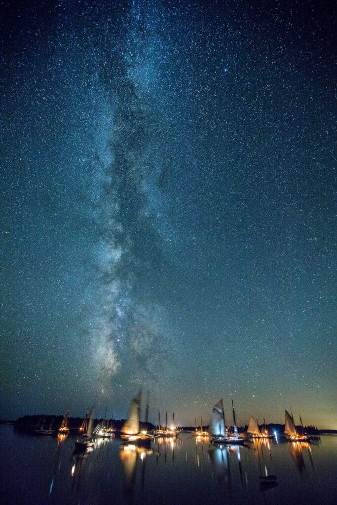 Sailboats anchored on calm water under a starry night sky with the Milky Way visible.