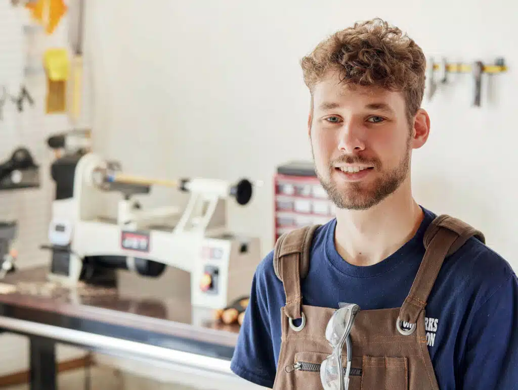 A smiling man wearing a workshop apron stands in front of a wood lathe.