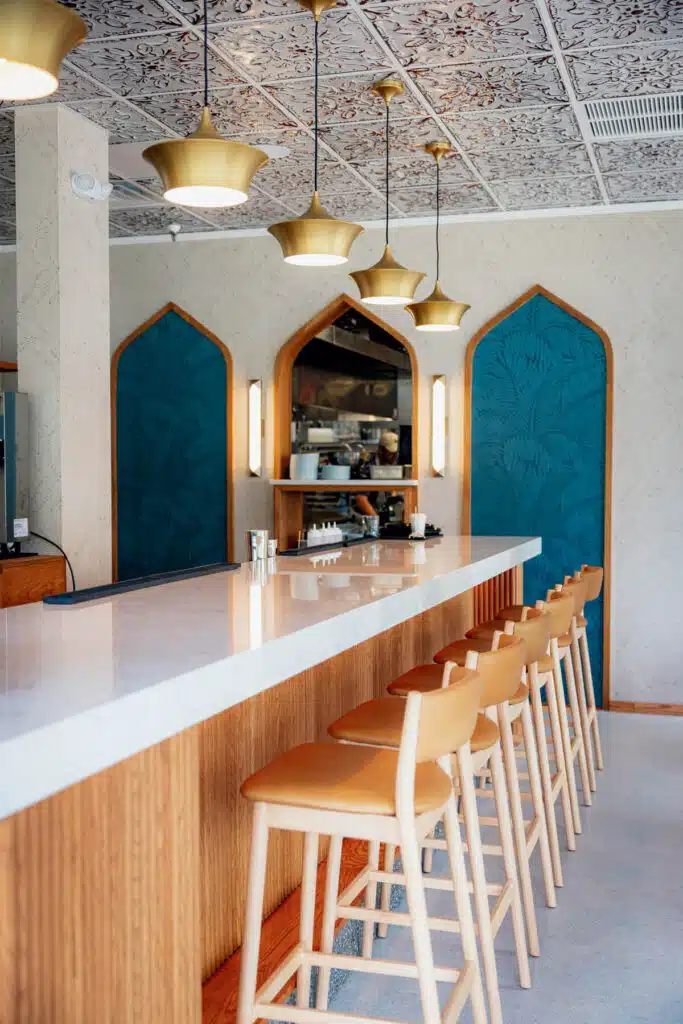A marble-topped bar with light wood stools, gold pendant lights, and arched teal panels on the back wall.