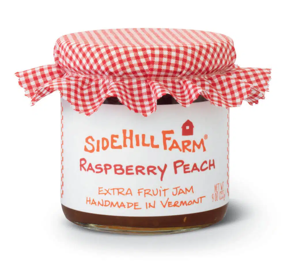 A jar of Sidehill Farm Raspberry Peach extra fruit jam with a red and white checkered cloth lid cover.