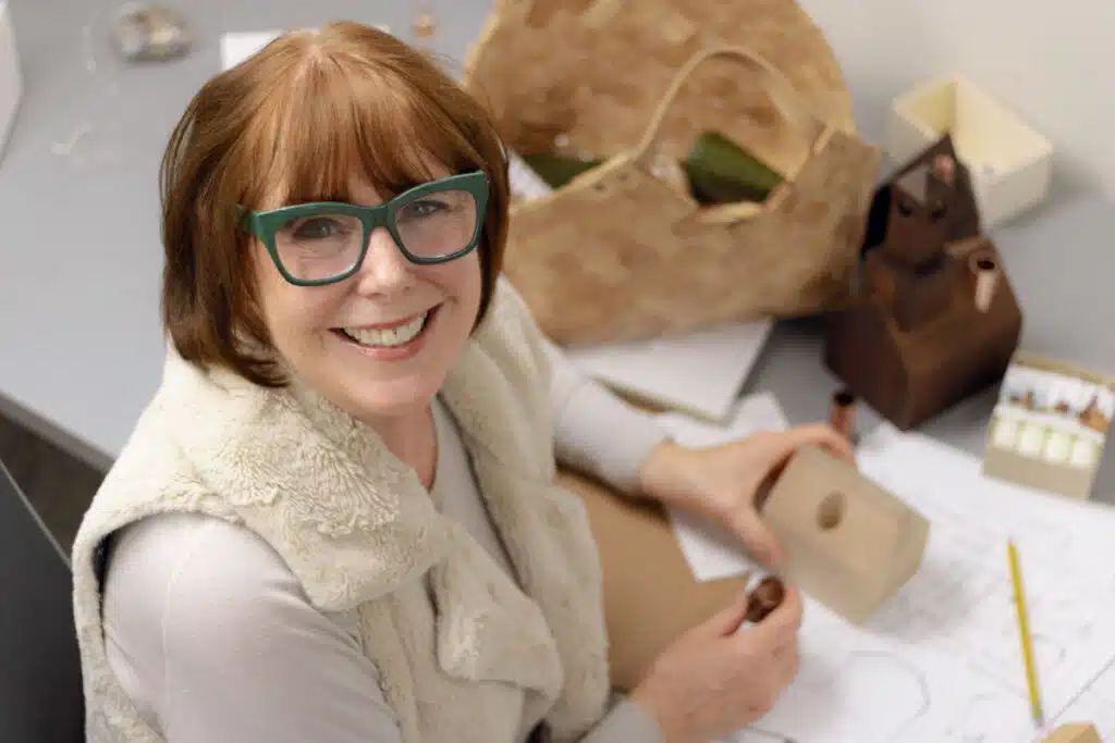 A smiling woman with glasses works at a desk with wooden craft pieces and a basket.