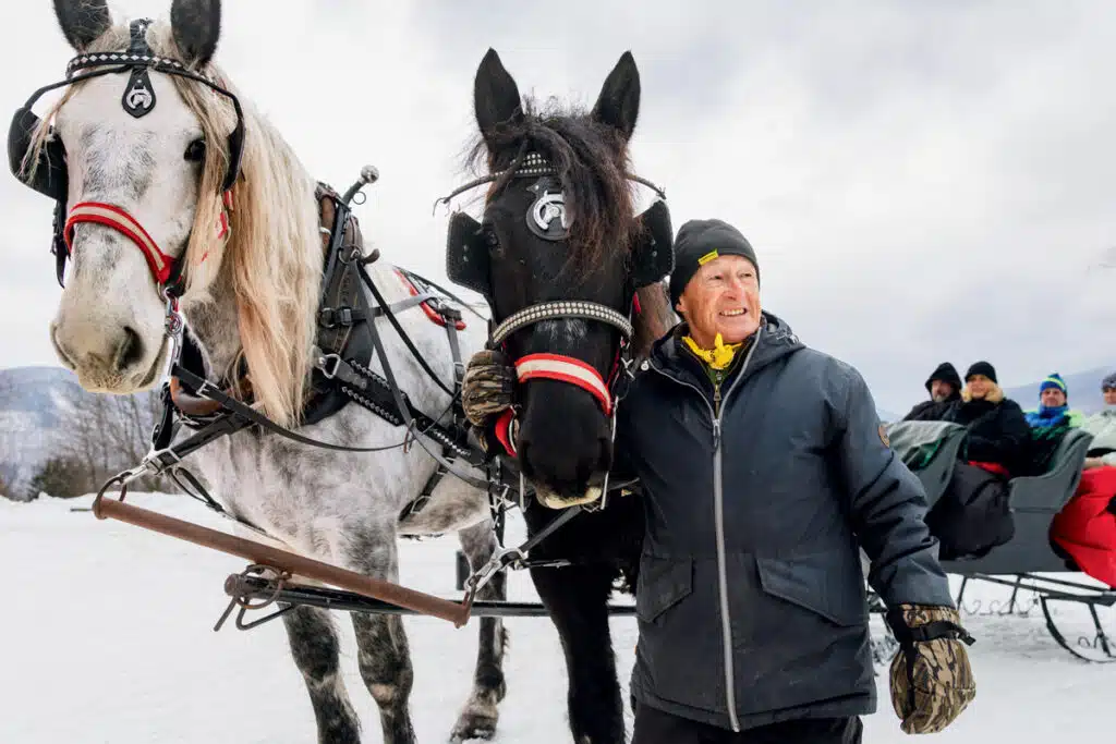 A man stands next to a team of horses hitched to a passenger sleigh in the snow.