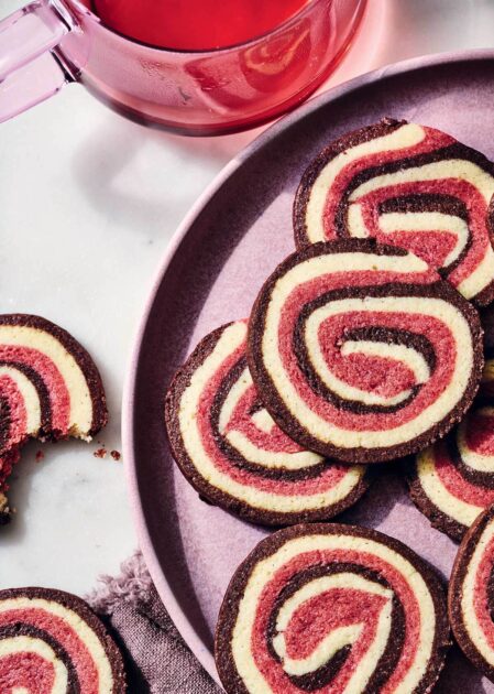Neapolitan Pinwheel Cookies. Red and cream-colored pinwheel cookies on a plate next to a glass teapot.